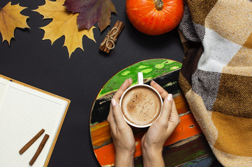 Cup of pumpkin cocoa or coffee in female hands, opened notebook, pumpkin, cinnamon, yellow autumn maple leaf, checkered plaid on black background top view copy space. Autumn flat lay background
