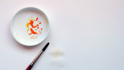 Flat lay of water color brush and a mixing tray with orange paint, on a white paper background