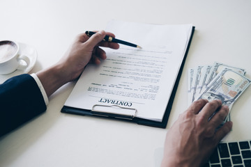 Man's hand in suit giving cash money and using pen point to signing a mortgage loan agreement in office, Mortgage and Home Loan concept.