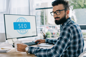 young successful seo manager sitting at workplace with computer and looking at camera
