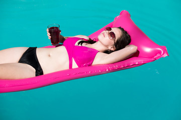 Beautiful woman floating on a bright pink raft in an outdoor swimming pool. Caucasian girl in bikini on holiday lounging in swimming pool in the sun on a pink raft. Resting, getaway, drink in hand.