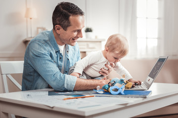 Working father. Cheerful delighted man playing with his son while working from home