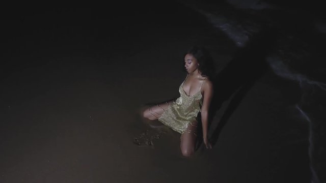 TRACKING SHOT Of A Beautiful Afro-American Girl In A Golden Dress Gracefully Posing On The Beach At Night