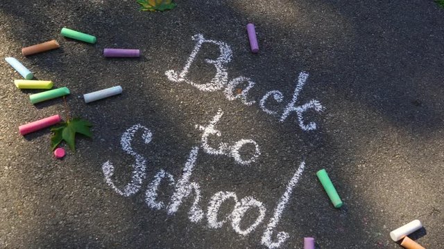 Back to school background. Words written by child outdoor on sunny  grey asphalt texture with white chalk. Many other colorful chalks laying around. Real time 4 k video footage.