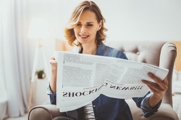 Calmness at home. Pleased young woman sitting on her armchair and reading newspaper