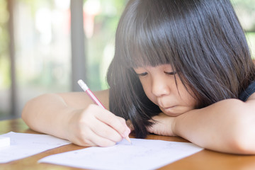Portrait of adorable Asia kid girl doing boring homework with bored feeling at home. Female cute child concentrate write on paper. Asian students practice math subject for test. Student study at house