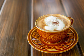 The  white ceramic cup of hot capucchino coffee on the wooden table in the restaurant or coffee shop with blur people at the background in thw warm yellow light and flare. 