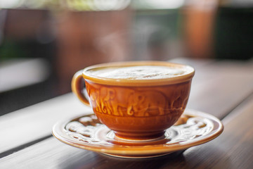 The  brown ceramic cup of hot capucchino coffee on the wooden table in the restaurant or coffee shop with blur people at the background in thw warm yellow light and flare. 