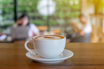 The  white ceramic cup of hot capucchino coffee on the wooden table in the restaurant or coffee shop with blur people at the background in thw warm yellow light and flare. 