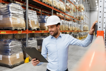 logistic business, shipment and people concept - businessman in helmet with clipboard checking goods at warehouse