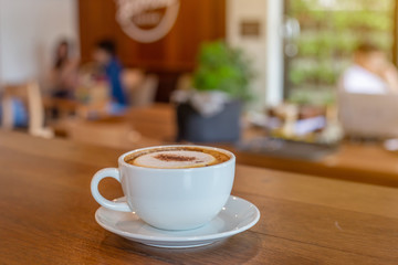 The  white ceramic cup of hot capucchino coffee on the wooden table in the restaurant or coffee shop with blur people at the background in thw warm yellow light and flare. 