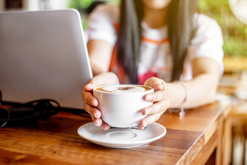 The  white ceramic cup of hot capucchino coffee on the wooden table in the restaurant or coffee shop with blur people at the background in thw warm yellow light and flare. 
