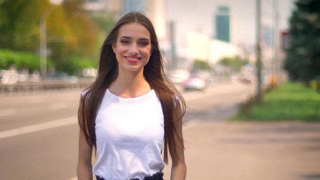 Sexy woman is standing on bus stop in daytime in summer, watching at camera, smiling, blurred road on background