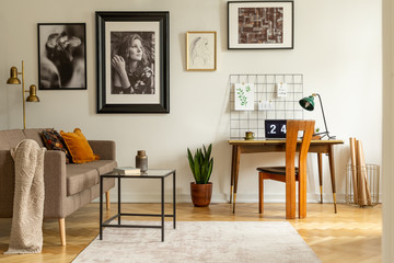 Sofa and table in open space interior with posters above desk with lamp and laptop. Real photo