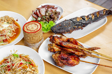 The set of Thai food such as papaya salad , grilled chicken , grilles meat , cat fish and sticky rice in the bamboo container on the wooden table. 