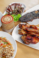 The set of Thai food such as papaya salad , grilled chicken , grilles meat , cat fish and sticky rice in the bamboo container on the wooden table. 