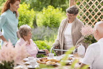 A group of pensioners with their caregiver having breakfast by a table on a porch outside a nursing home.
