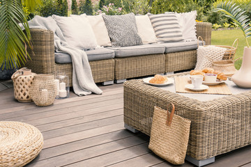 Wicker patio set with beige cushions standing on a wooden board deck. Breakfast on a table on a backyard porch. - obrazy, fototapety, plakaty