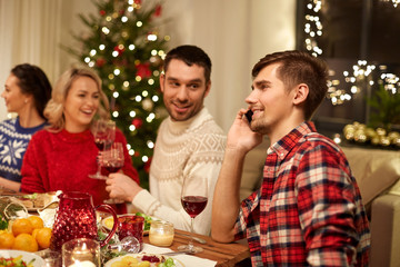 holidays, communication and celebration concept - happy young man calling on smartphone and having christmas dinner with friends at home