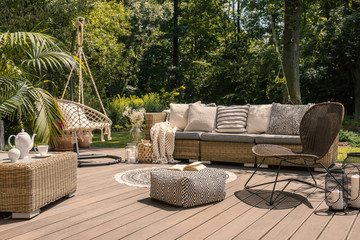 A rattan patio set including a sofa, a table and a chair on a wooden deck in the sunny garden. - obrazy, fototapety, plakaty