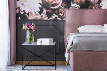 Flowers on black table next to pink and grey bed in bedroom interior with wallpaper. Real photo