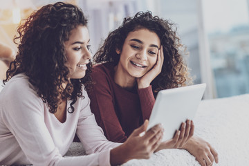 Relax on weekend. Happy sisters lying on bed together while looking at their gadget with pleasure