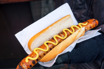 Person's Gloved Hand Holding Bratwurst or Rostbratwurst, a Traditional German Sausage, with Mustard on a Bun