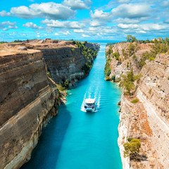 Beautiful scenery of the Corinth Canal in a bright sunny day against a blue sky with white clouds. Among the rocks floating white ship in turquoise water. - obrazy, fototapety, plakaty