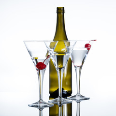 Three martini glasses filled with a clear drink in front of a brown bottle against a white background. Two red cherries in two of the glasses