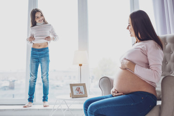 Whos bigger. Positive pregnant mother and her little cute daughter looking at each other while...