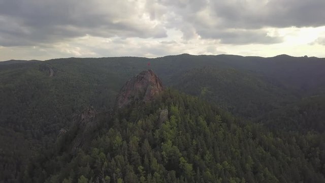  Aerial Drone Footage View: Aerial View flight over flight of high mountains covered with green coniferous pine trees and cedars. At the top of the mountain is a huge stone in the flag, set in honor o