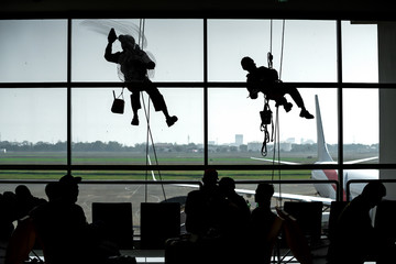 Employees are using a rope to rappelled down to clean the glass of an airport building. There are passengers waiting to board and are looking at the cleaning staff. Silhouette concept.