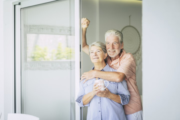 cheerful adult couple at home looking outside together with joy and peace. happy retired caucasian man and woman enjoying his new lifestyle with love and peaceful time. hug and relationship together