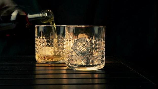 barman pouring whiskey in the two glasses with ice cubes on wood table and black dark background, focus on ice cubes, whisky relax time on warm atmosphere