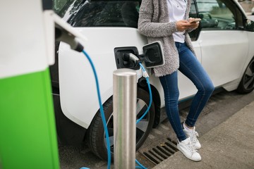 Woman using mobile phone while charging electric car at charging