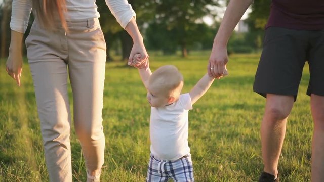 Male infant toddler learning to walk in the park with his parents. Mother and father holding hands of their son, aking steps on the green grass. Outdoors. Slow motion.