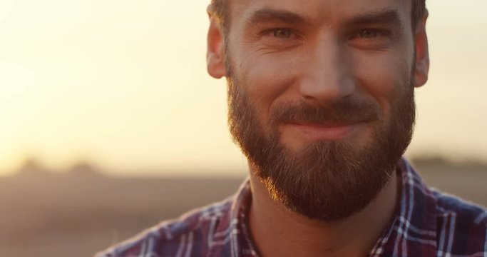 Close up of the Caucasian good looking young man with a beard smiling to the camera and then looking down early in the morning at his field. Portrait.