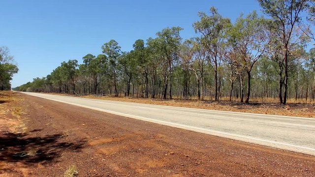 White heavy truck with two trailers passing by empty road in Outback Australia. Desert freeway flanked by trees in Northern Terrotory rural area. Long distance goods transportation concepts