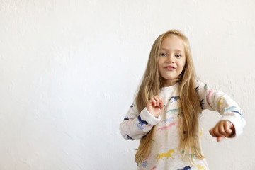 Obraz na płótnie Canvas Little blonde girl with long golden hair dancing, smling and having fun over white textured plaster wall background. Five years old blonde female child posing. Copy space for text.