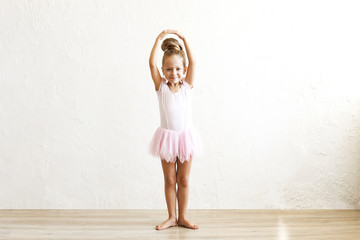 Little blonde balerina girl dancing and posing in dance club with wooden floot an white textured plaster wall. Young ballet dancer in pink tutu dress, having fun and smiling. Backgroud, copy space. © Evrymmnt