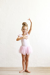 Little blonde balerina girl dancing and posing in dance club with wooden floot an white textured...
