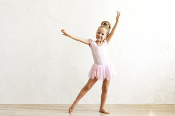 Little blonde balerina girl dancing and posing in dance club with wooden floot an white textured plaster wall. Young ballet dancer in pink tutu dress, having fun and smiling. Backgroud, copy space.