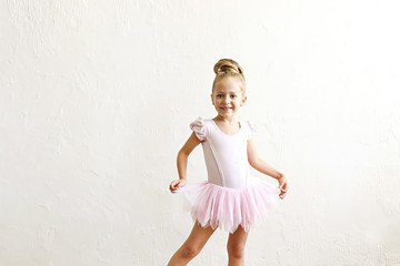 Little blonde balerina girl dancing and posing in dance club with wooden floot an white textured plaster wall. Young ballet dancer in pink tutu dress, having fun and smiling. Backgroud, copy space. © Evrymmnt