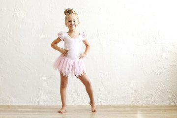 Little blonde balerina girl dancing and posing in dance club with wooden floot an white textured...