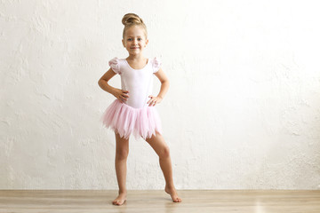 Little blonde balerina girl dancing and posing in dance club with wooden floot an white textured plaster wall. Young ballet dancer in pink tutu dress, having fun and smiling. Backgroud, copy space.