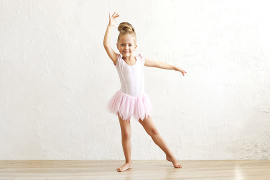 Little blonde balerina girl dancing and posing in dance club with wooden floot an white textured plaster wall. Young ballet dancer in pink tutu dress, having fun and smiling. Backgroud, copy space.
