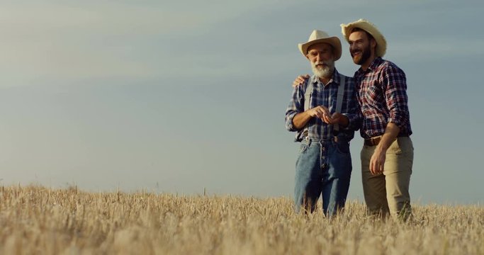 Handsome young farmer in a hat standing together with his old father in the field during harvest time, talking and showing something by pointing with a finger.