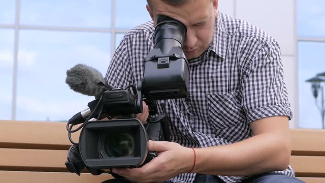 The cameraman sitting on a bench near the building is videotaping in a TV camera