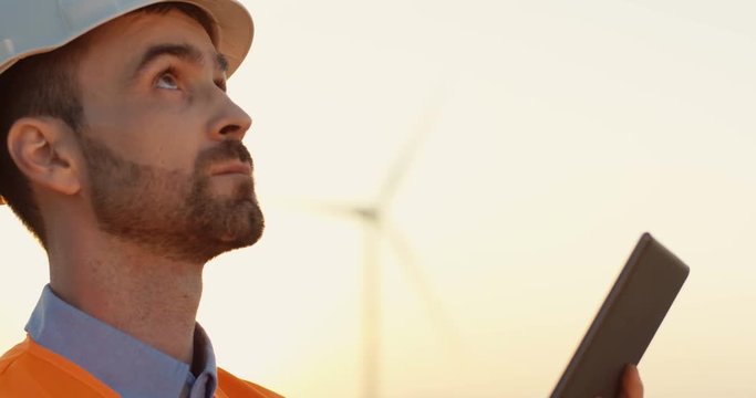 Close up of the handsome Caucasian man in the helmet checking something on the tablet computer and looking up while working outside.