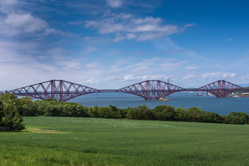 Fototapeta na wymiar Queensferry, Scotland, UK - June 14, 2012: Red metal iconic Forth Bridge for trains over Firth of Forth under blue sky. Green field up front.